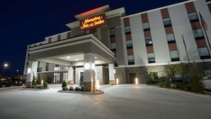 A Hampton Inn & Suites hotel entrance with a modern design, featuring a prominent sign, a well-lit driveway, and a landscaped area with small trees and shrubs.