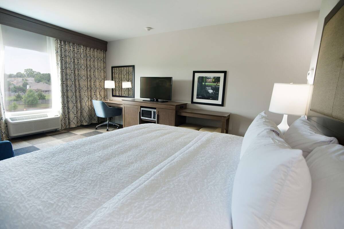 A hotel room with a large bed, a desk with a television, a chair, and a window with curtains.