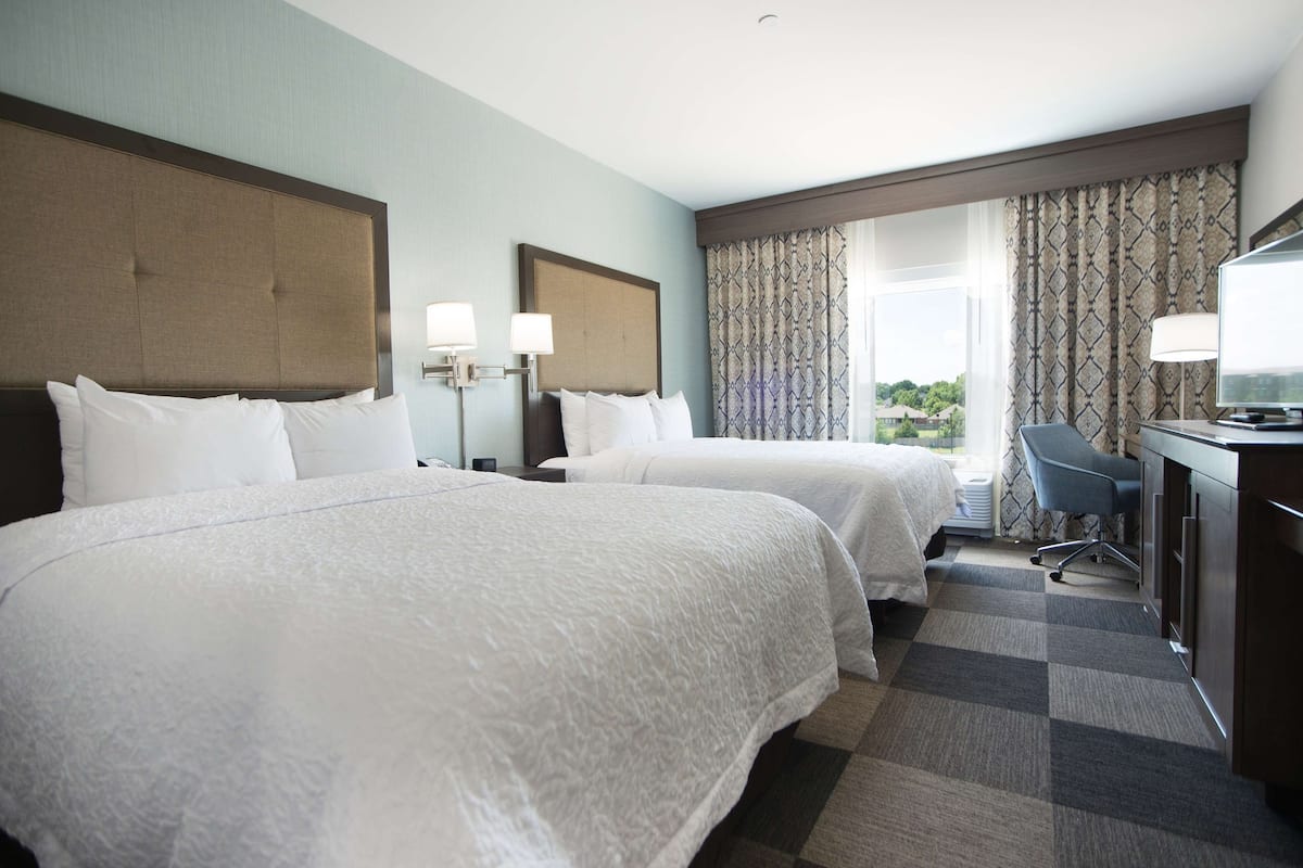 A hotel room with two beds, a desk, a chair, and a view of the outdoors.
