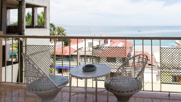 Een balkon met twee rieten stoelen en een kleine tafel, met uitzicht op een strand en gebouwen.