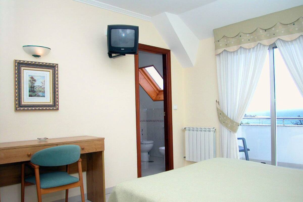 Habitación de hotel con cama, escritorio con silla, televisor, un cuadro enmarcado y vista al mar por la ventana.