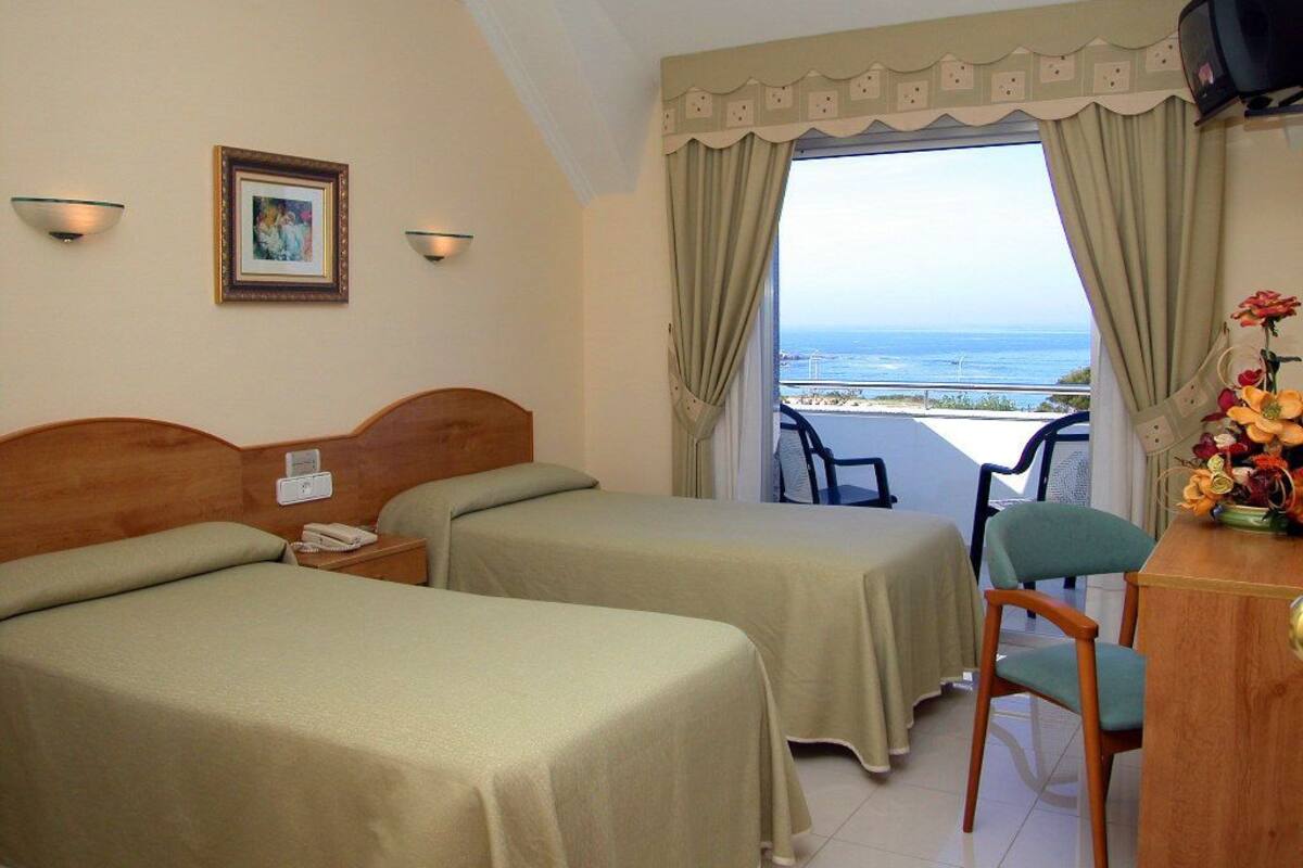 Habitación de hotel con dos camas, un escritorio, una silla, un televisor y un balcón con vista al mar.