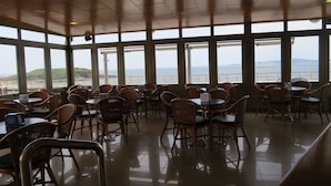 Un restaurante con amplios ventanales que ofrece vistas al mar y a las colinas. Mesas y sillas de mimbre están dispuestas para comer.