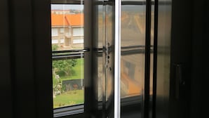 Un ascensor con acabado metálico y puerta de vidrio, que da a un balcón con vistas a edificios y follaje.