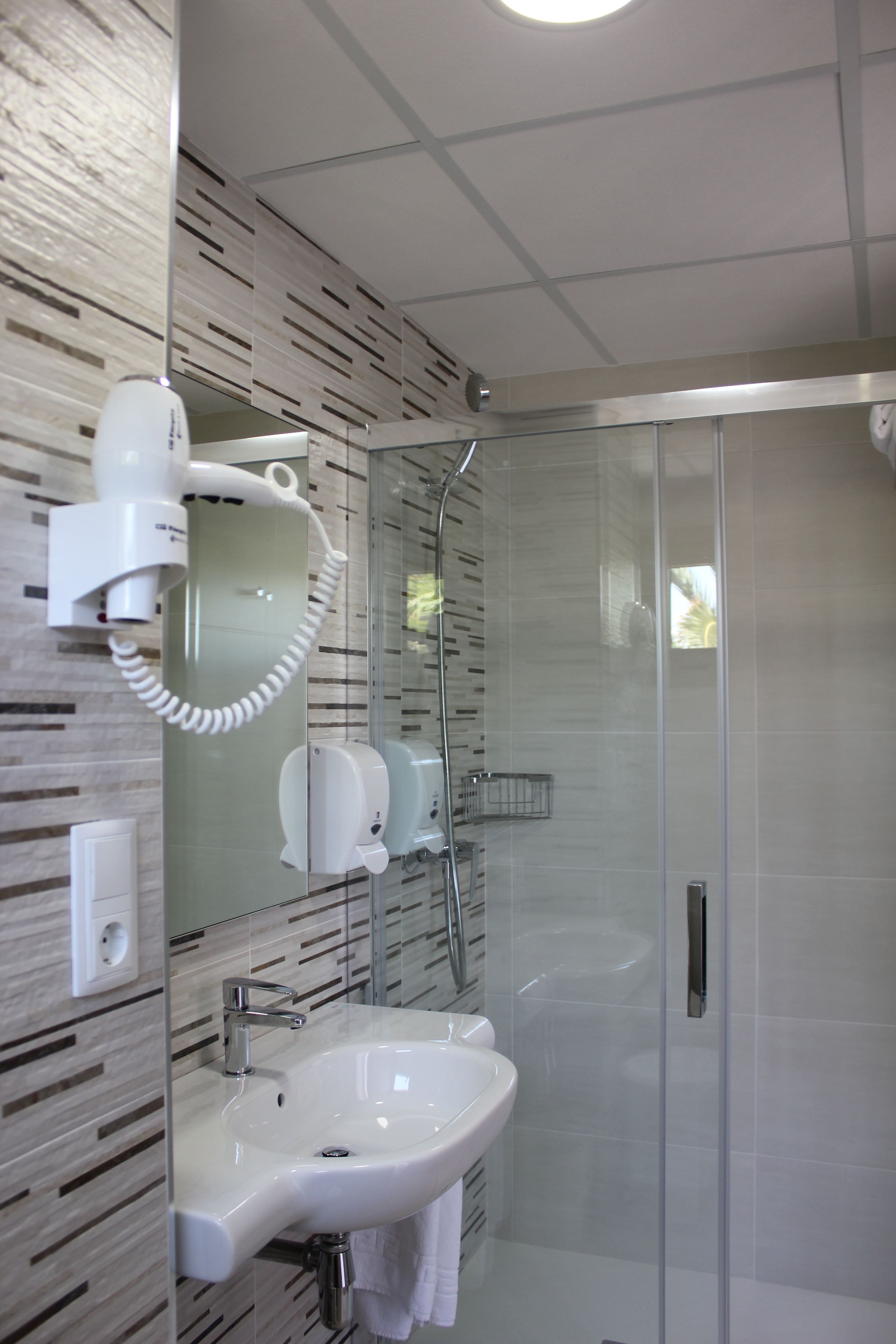Un baño moderno con cabina de ducha de vidrio, un lavamanos blanco y un secador de pelo fijado a la pared.