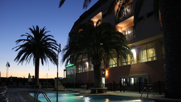 Zona de piscina con sillones de descanso y una palmera, rodeada por un edificio con balcones y ventanas.