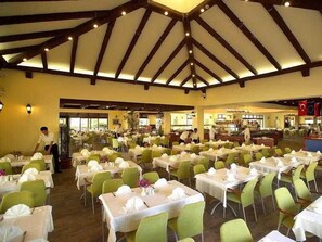 A large banquet hall with tables set for dining, a high ceiling with wooden beams, and a view of the kitchen area in the background. - Gypsophila Holiday Village (Alanya)