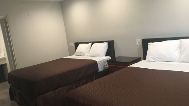 A hotel room with two single beds, each with a brown bedspread and white pillows. The room has a simple layout with a nightstand between the beds and a small table.
