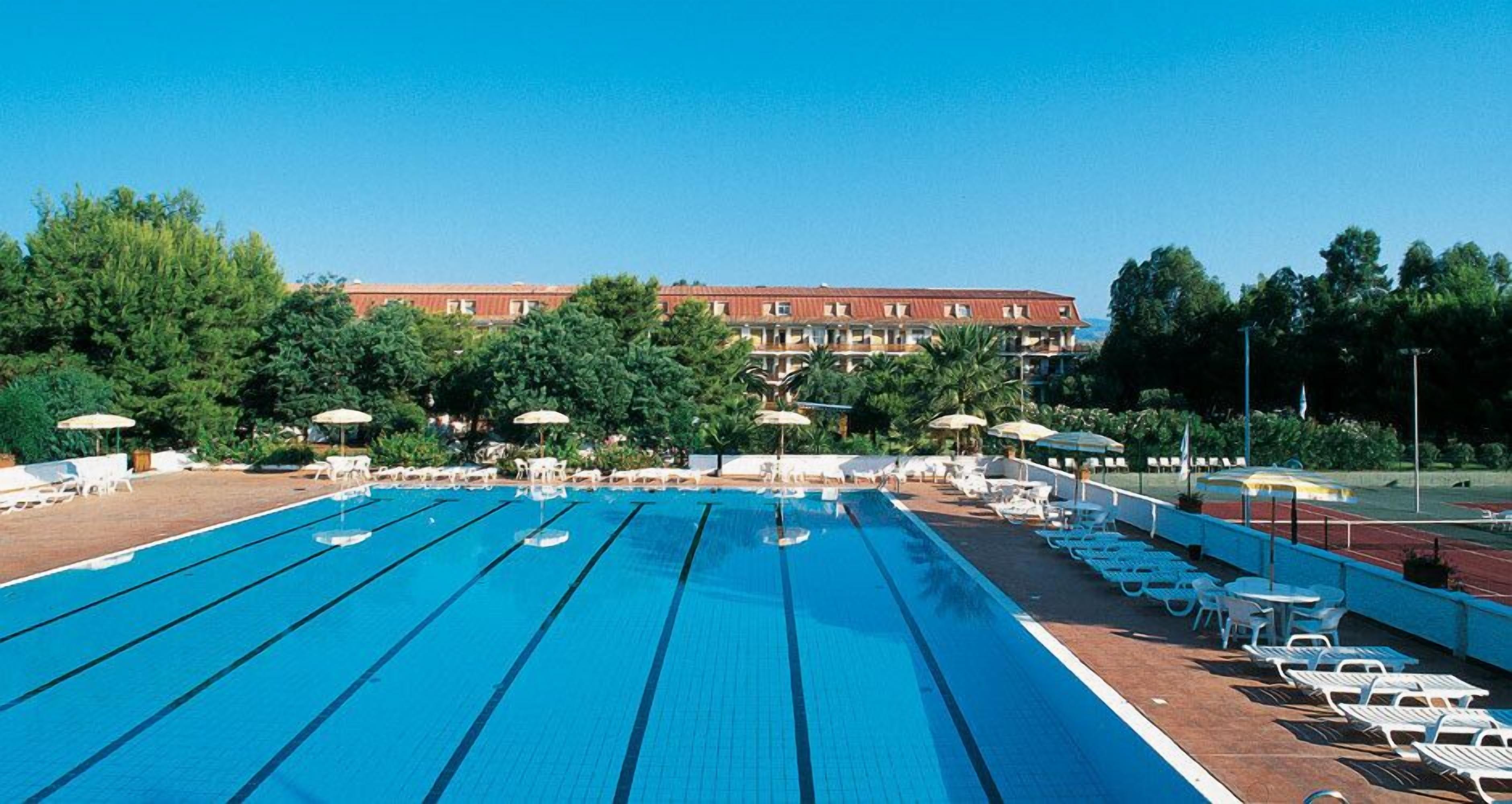 A swimming pool with multiple lanes, sun loungers, and umbrellas, surrounded by trees and a building in the background.