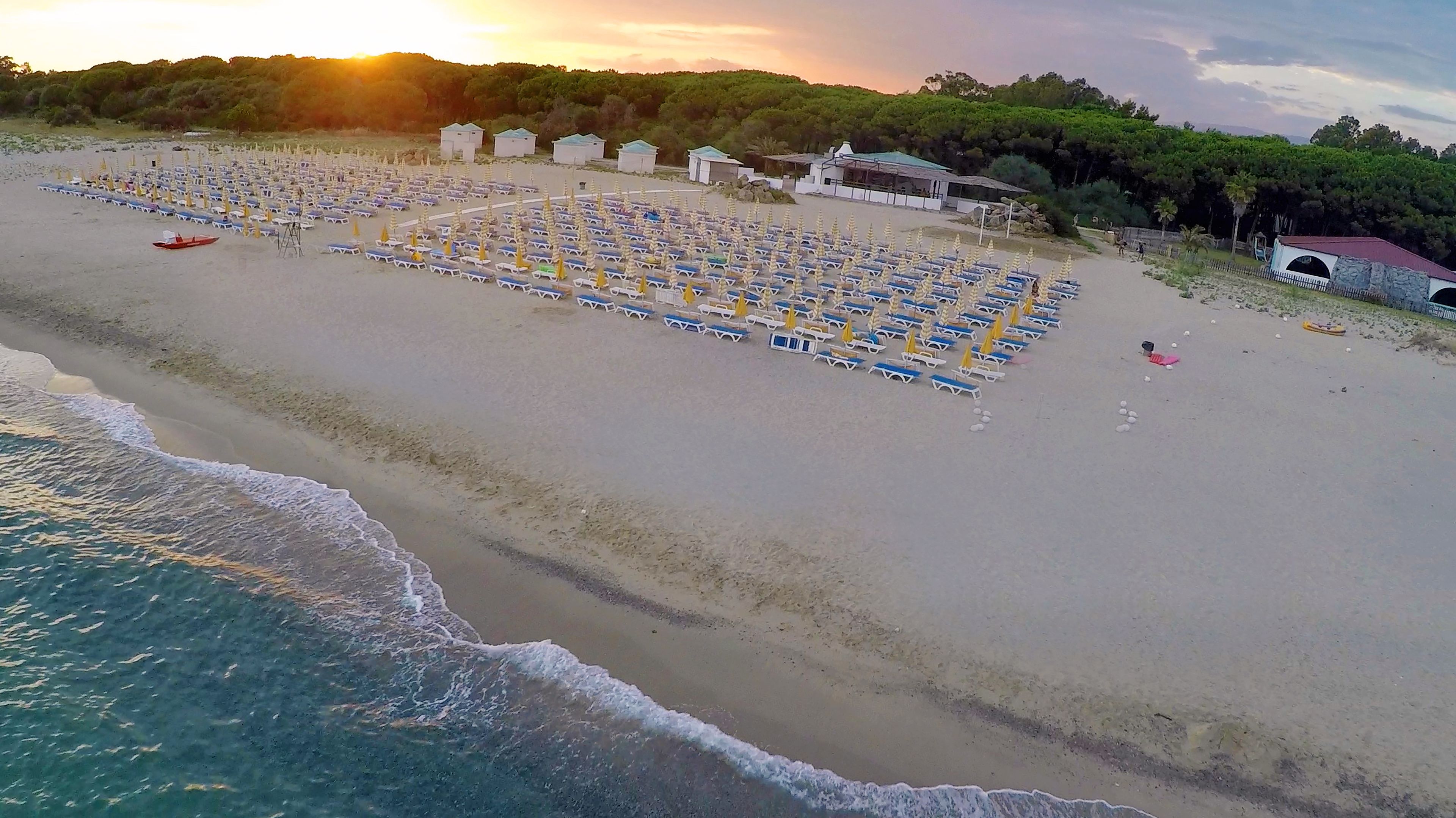 Una spiaggia con file di lettini e ombrelloni, una piccola imbarcazione e alcuni edifici sullo sfondo.