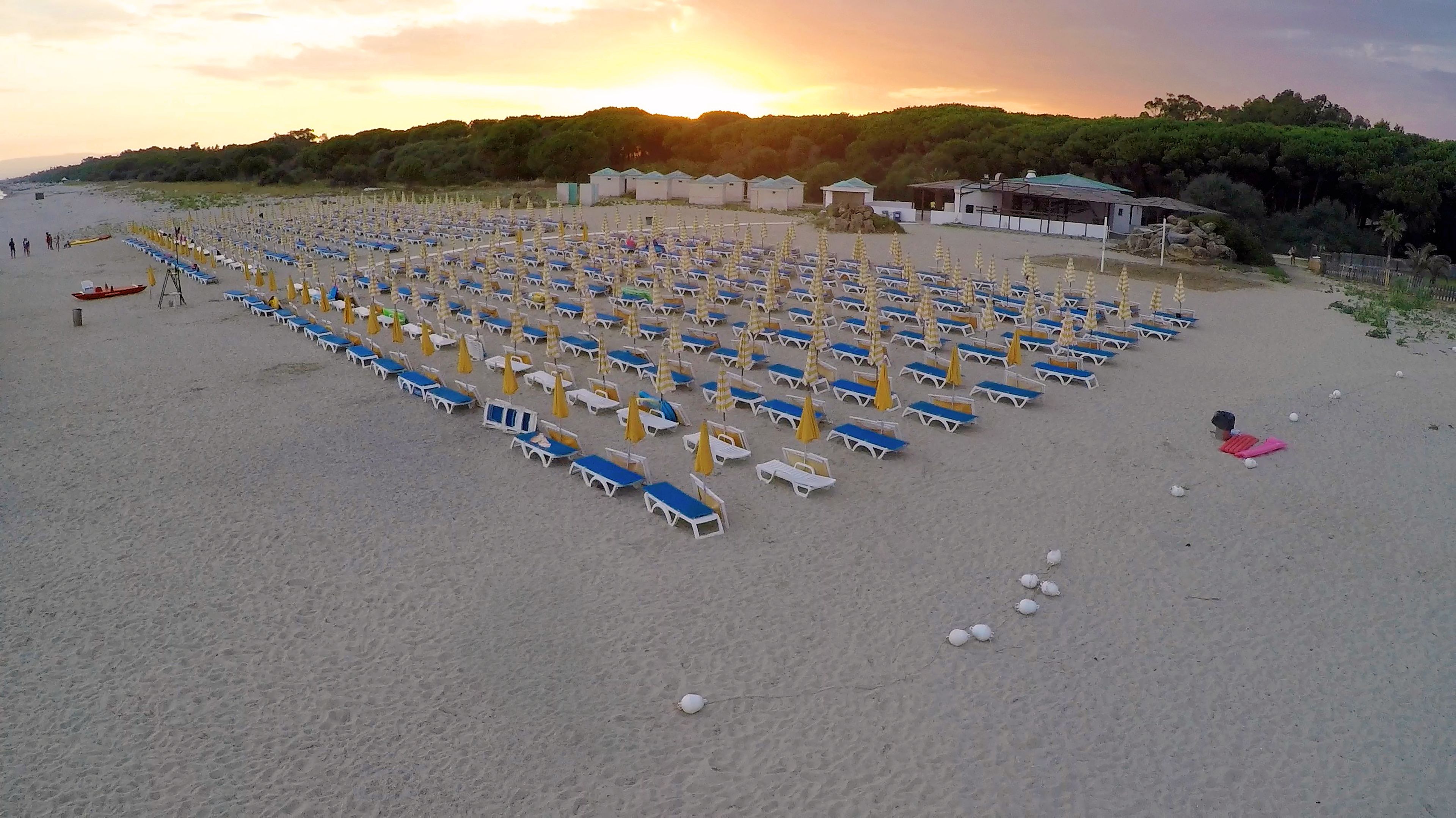 Ein Strand mit Reihen von Liegestühlen und Sonnenschirmen, einigen Personen und Gebäuden im Hintergrund.