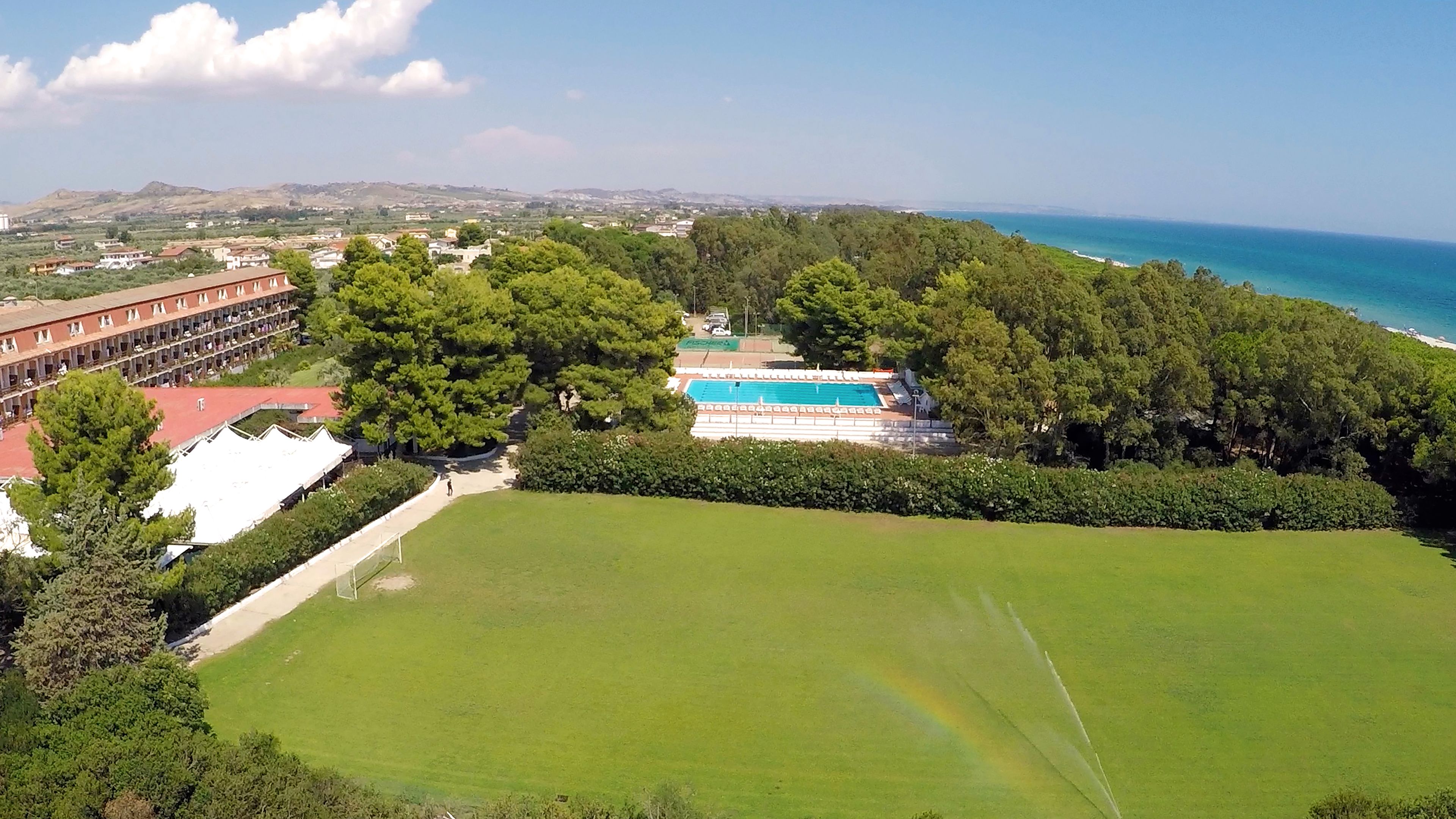 An aerial view of a hotel with a swimming pool, lush greenery, and a view of the sea.