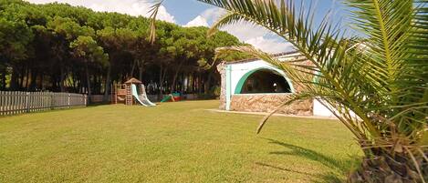A grassy park area with a playground, including a slide and climbing structures, and a small pavilion with a curved roof.