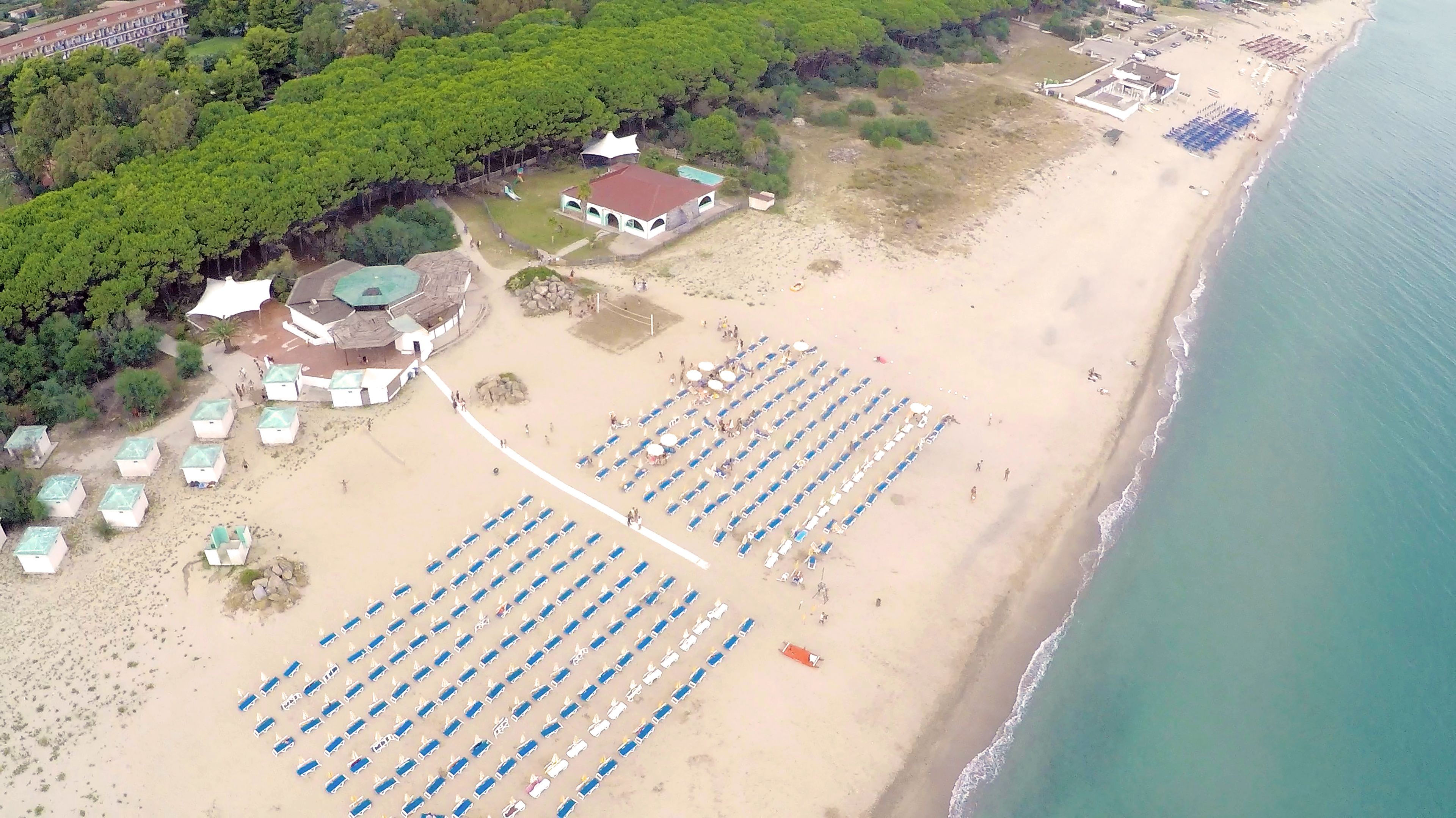 An aerial view of a beach with rows of blue beach chairs and umbrellas, a few small buildings, and a large body of water.