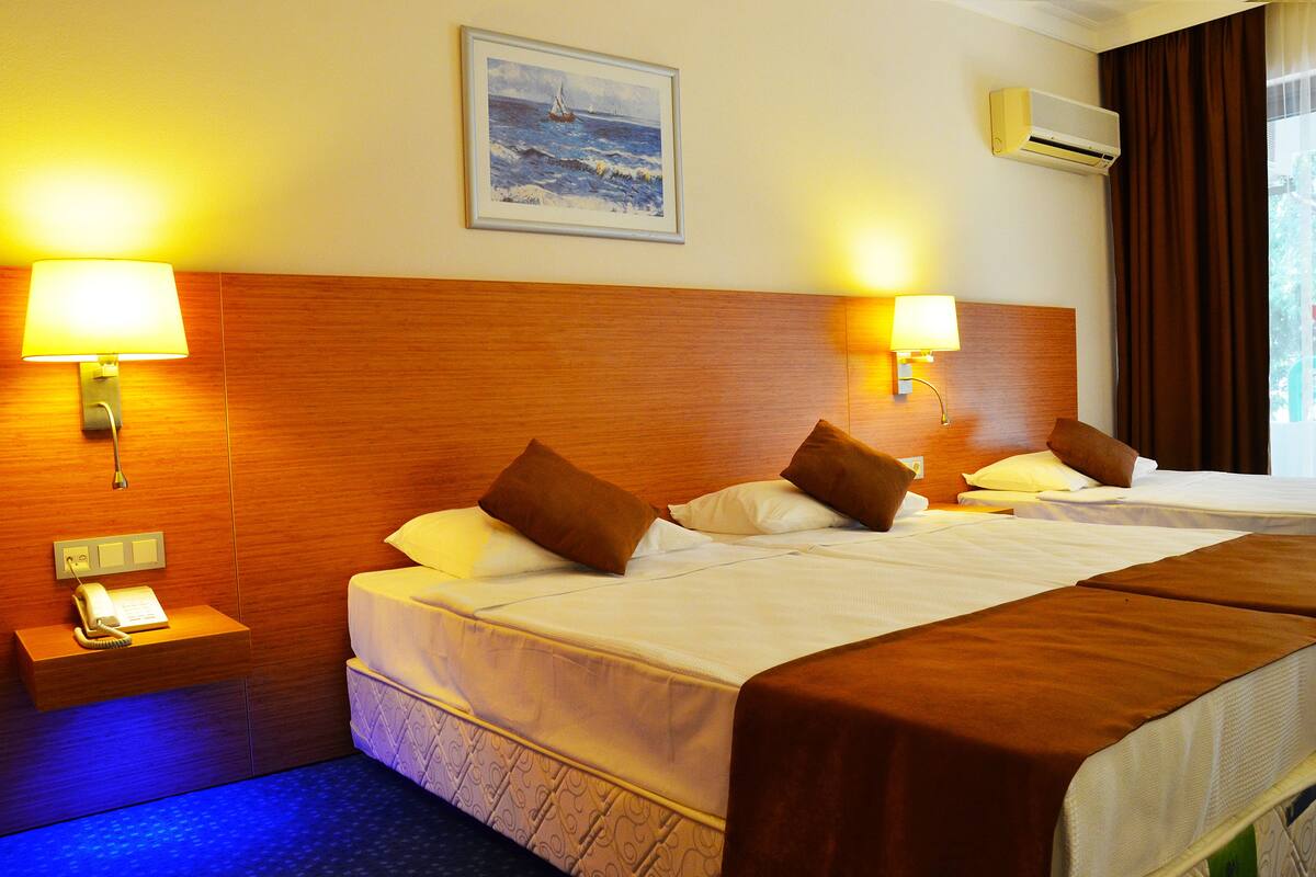 A hotel room with two beds, a wooden headboard, bedside lamps, a painting on the wall, and an air conditioning unit.
