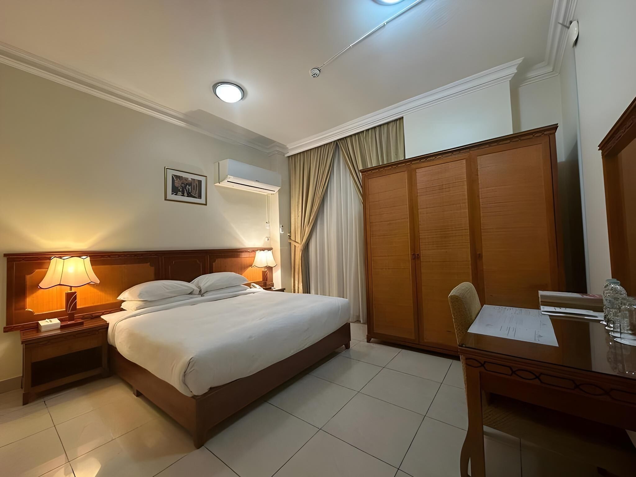 A hotel room with a bed, bedside lamps, a wardrobe, a desk with papers, and an air conditioning unit.