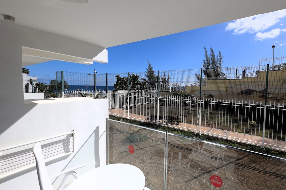 A balcony with a view of a fenced outdoor area, including a pool and a building with a yellow facade.