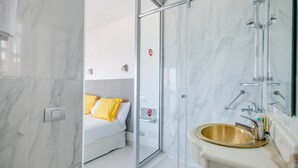 Un baño moderno con cabina de ducha de vidrio, lavamanos dorado y una cama con almohadones amarillos al fondo.