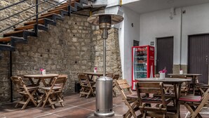 An outdoor patio area with wooden tables and chairs, a metal heater, a red Coca-Cola refrigerator, and a stone wall. - El Museo Hotel Boutique (La Paz)