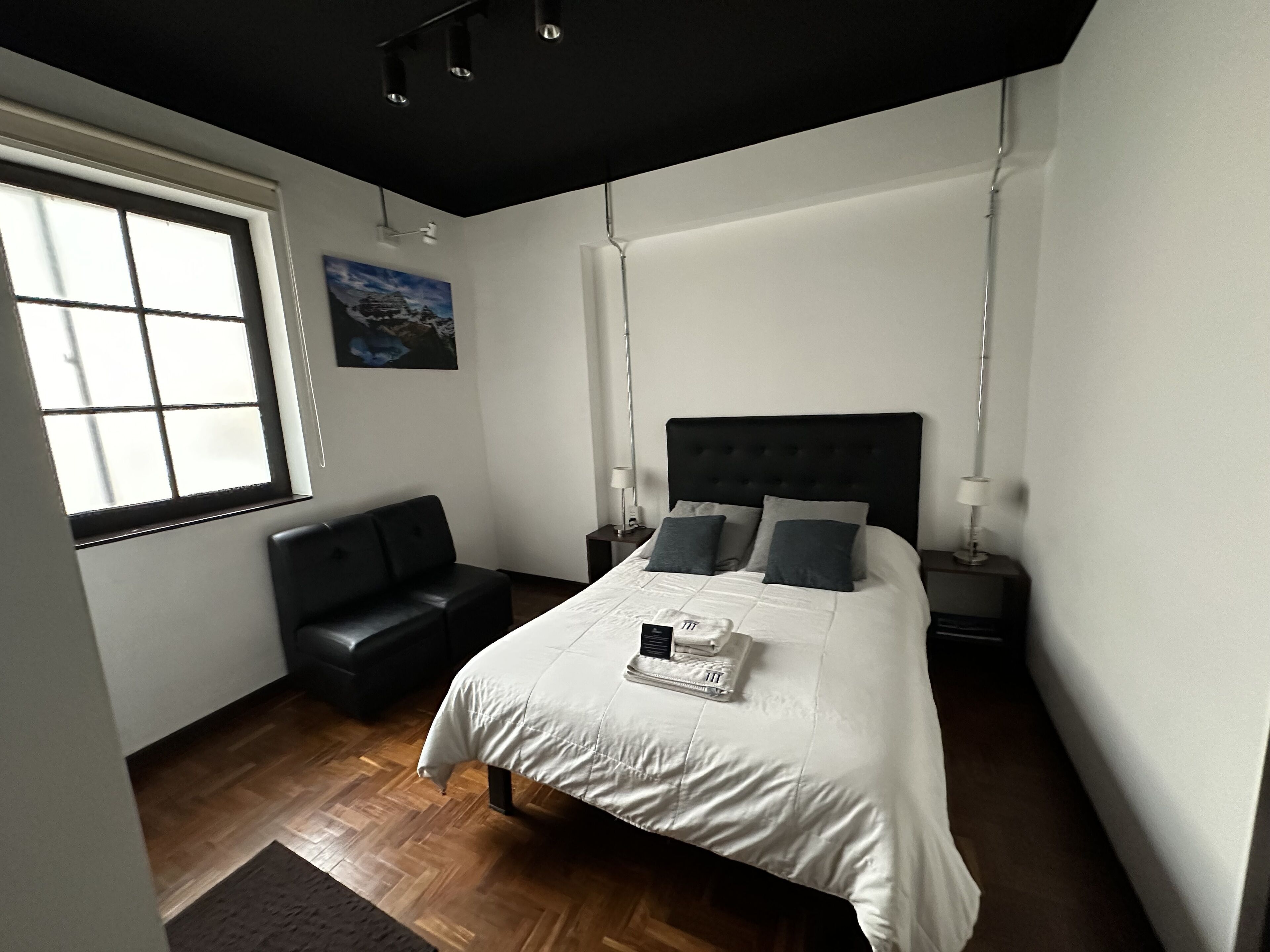 A modern bedroom with a large bed, a dark leather sofa, and a window providing natural light.