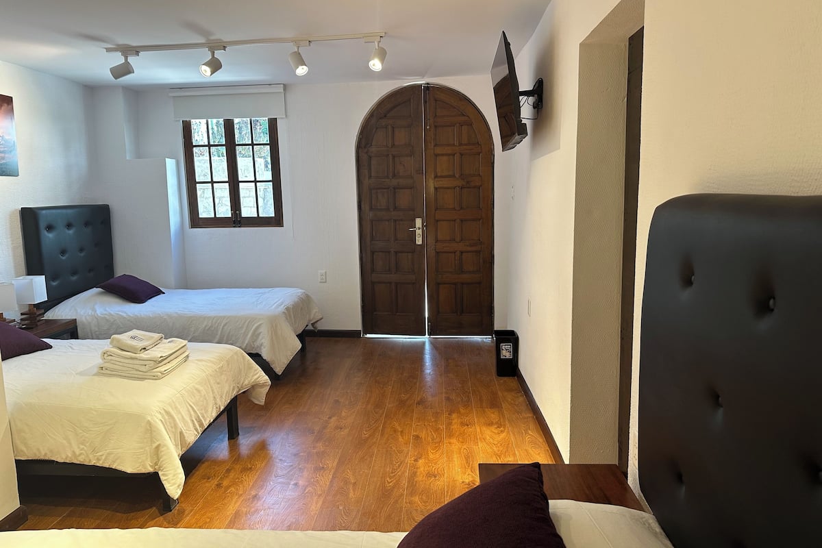 A hotel room with two beds, a wooden door, a TV mounted on the wall, and a window with blinds.