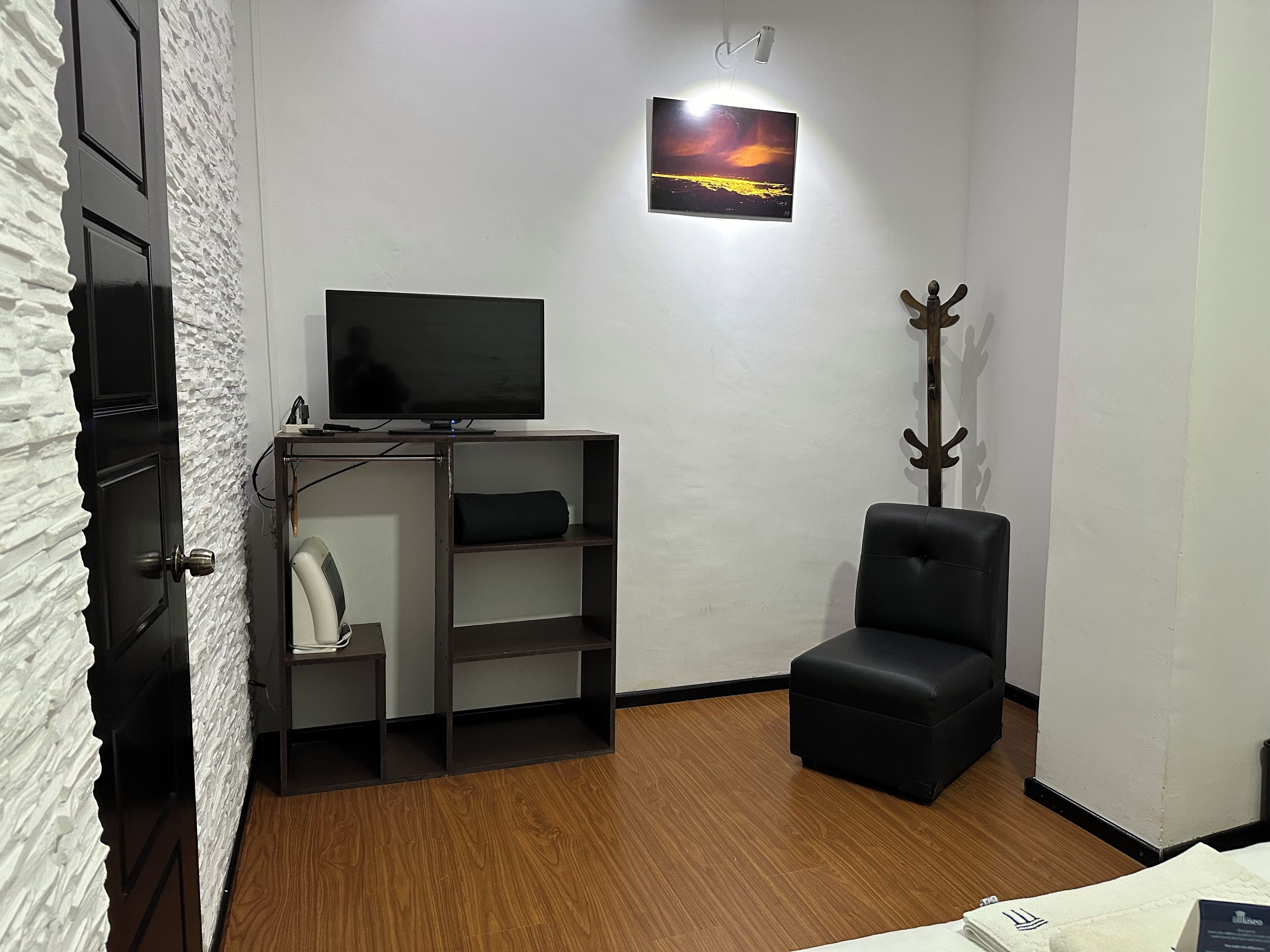 A small, modern room with a flat-screen TV on a wooden stand, a black chair, a coat rack, and a small shelf unit.
