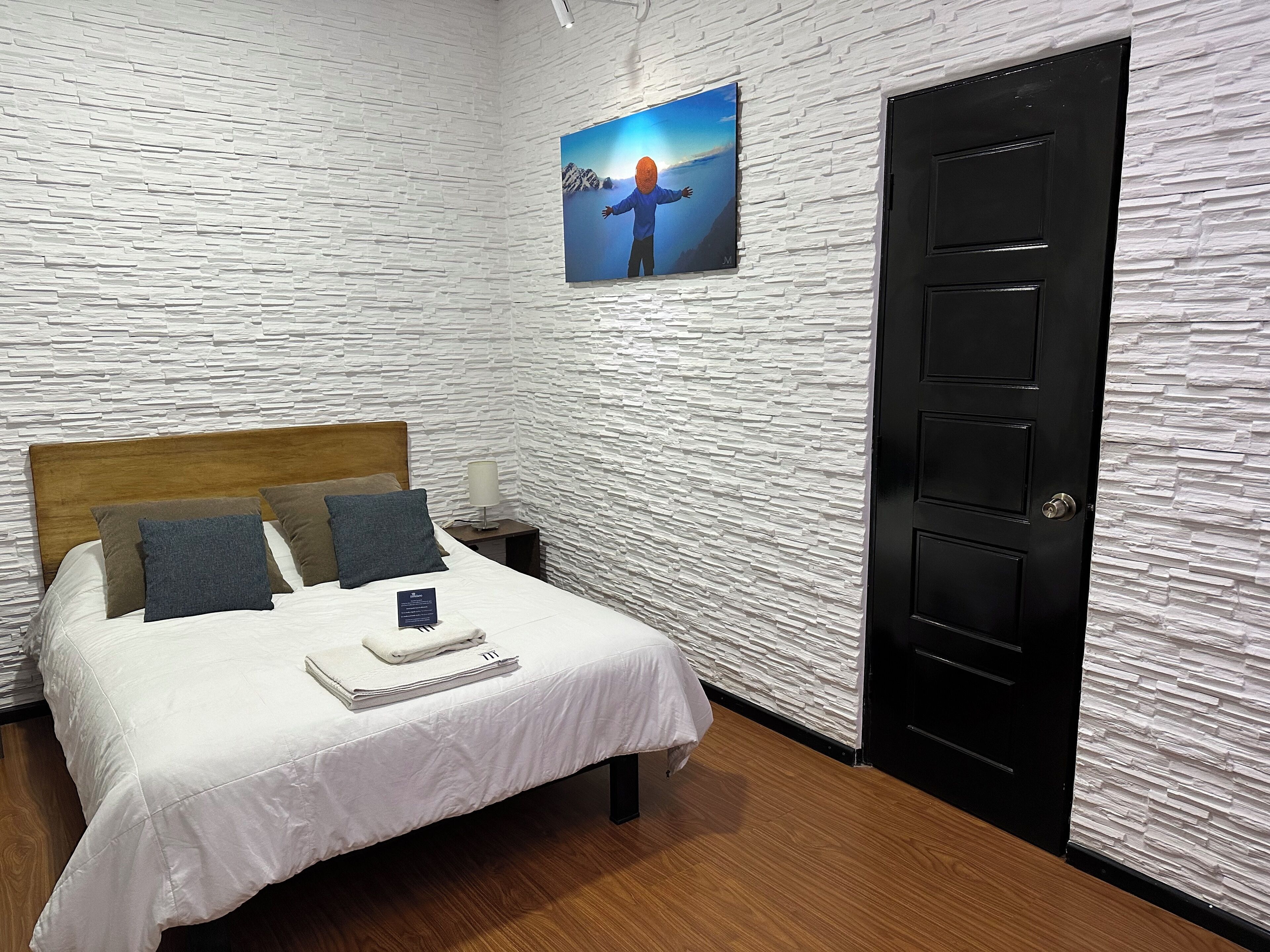 A bedroom with a white textured wall, a wooden headboard, a bed with white bedding, two pillows, a nightstand with a lamp, a framed picture on the wall, and a black door.