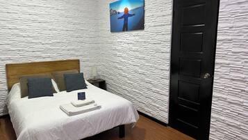 Un dormitorio con pared blanca texturizada, cabecera de madera, cama con ropa blanca, dos almohadas, mesita de noche con lámpara, un cuadro enmarcado en la pared y una puerta negra.