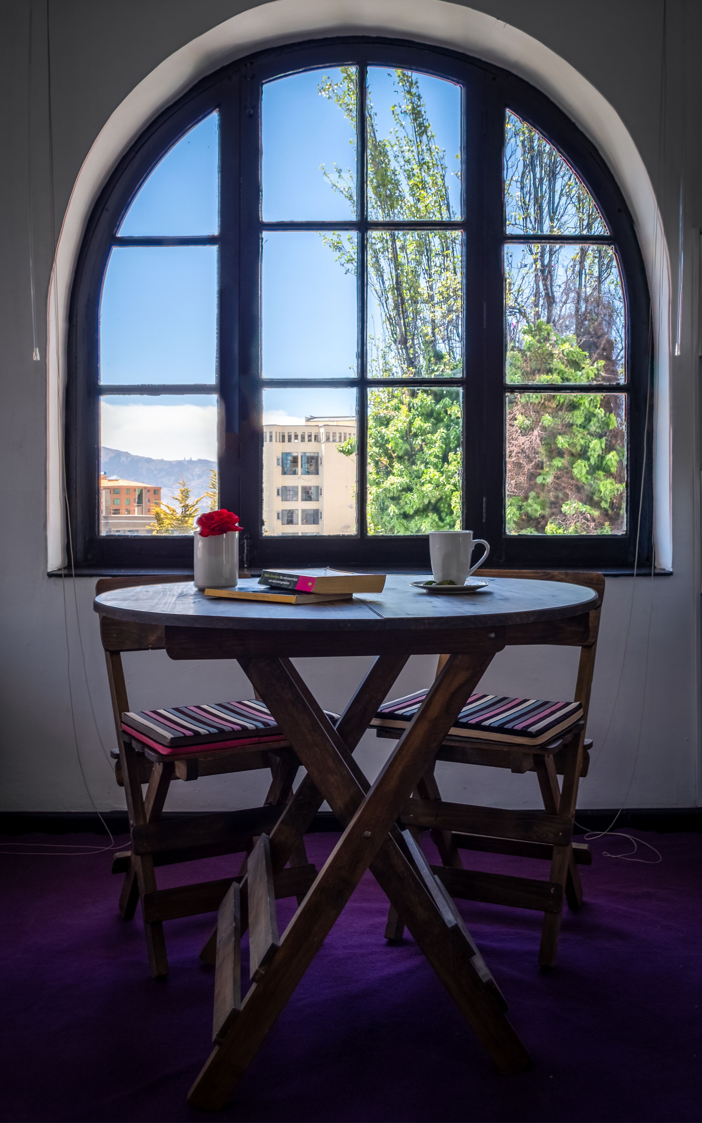 A room with a large arched window, a round table, two wooden chairs, a white vase with a red flower, and a white mug.