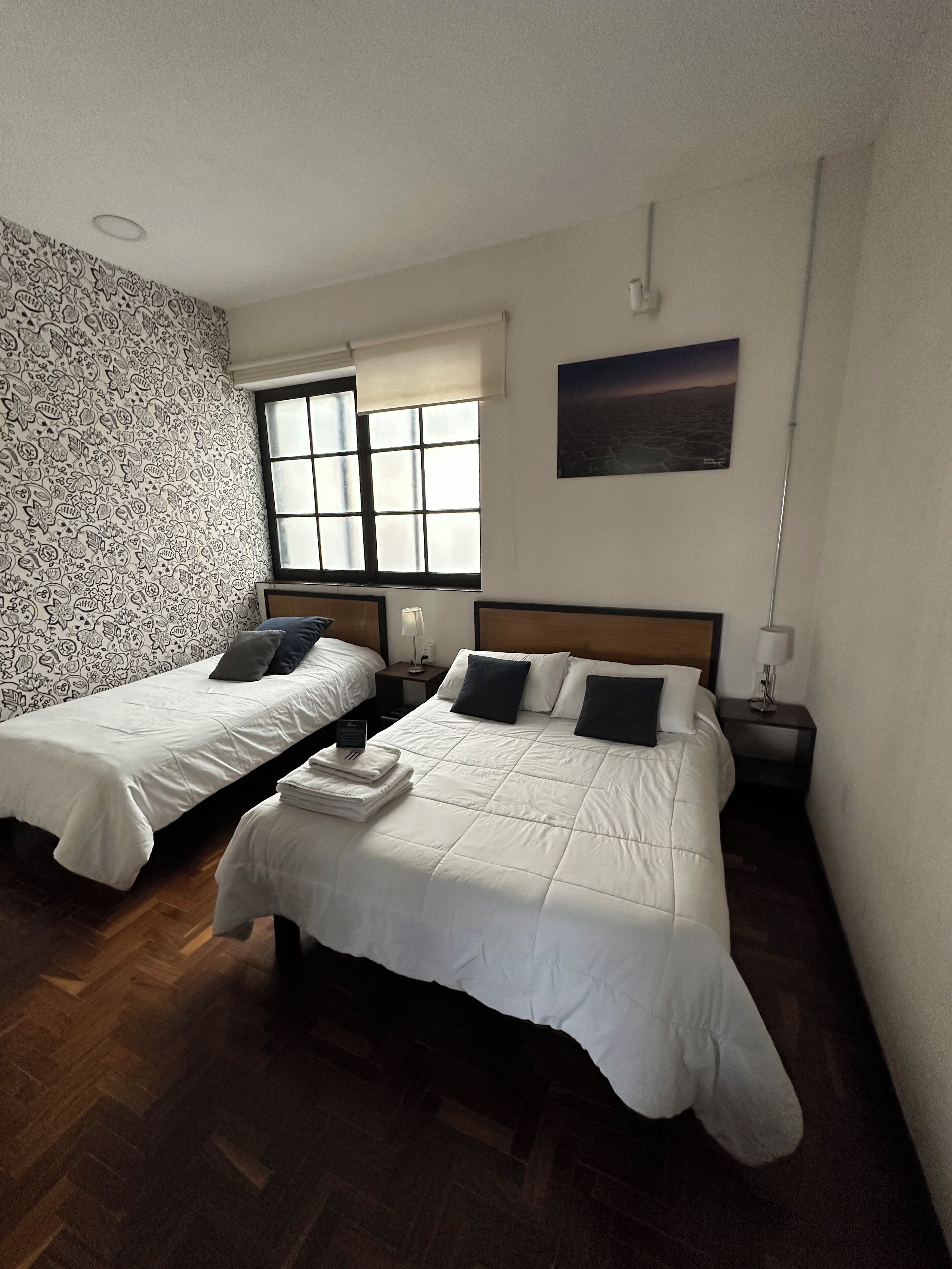 A hotel room with two beds, a patterned wall, a window with blinds, a nightstand with a lamp, and a painting on the wall.