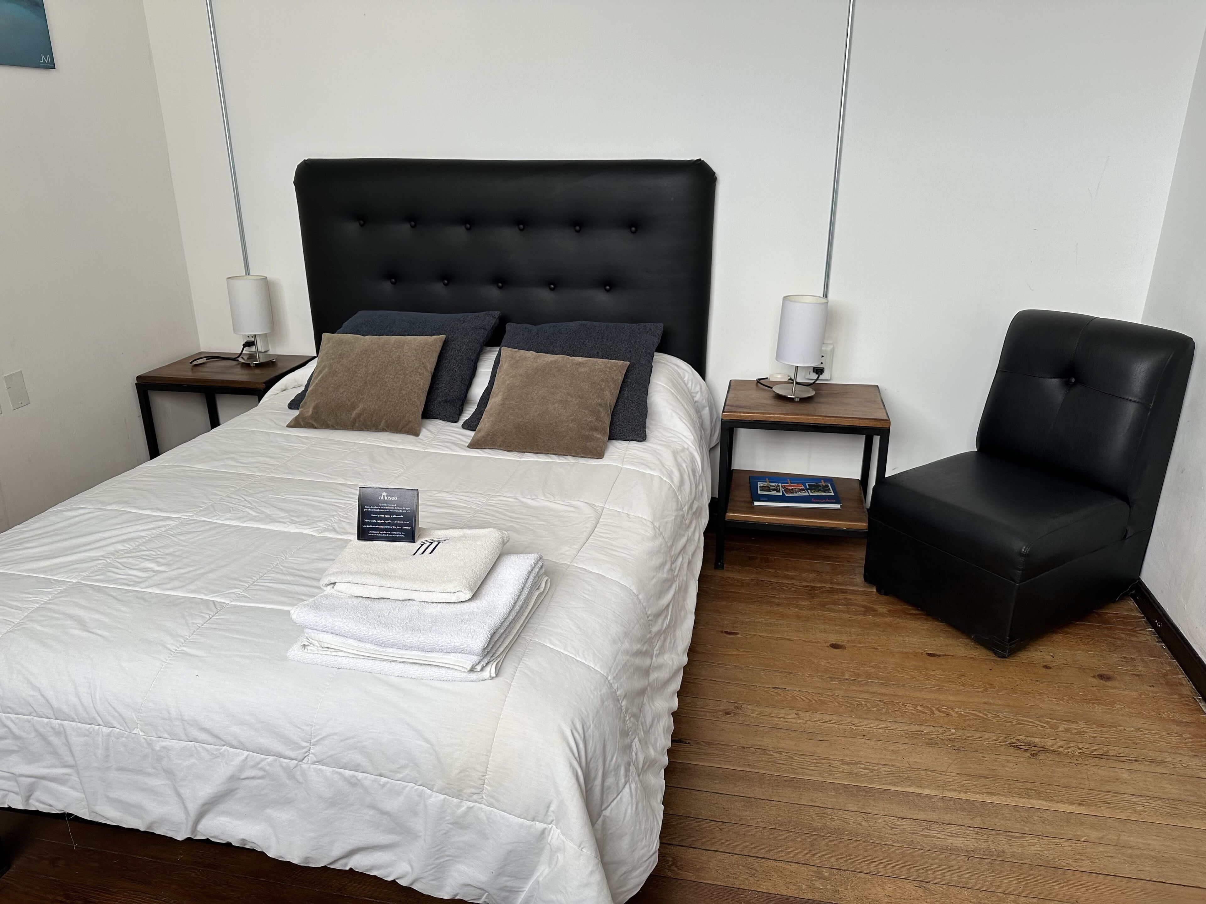 A neatly made bed with a white duvet, two pillows, and a folded towel. There are two bedside tables, each with a lamp and a book. A black leather chair is against the wall.