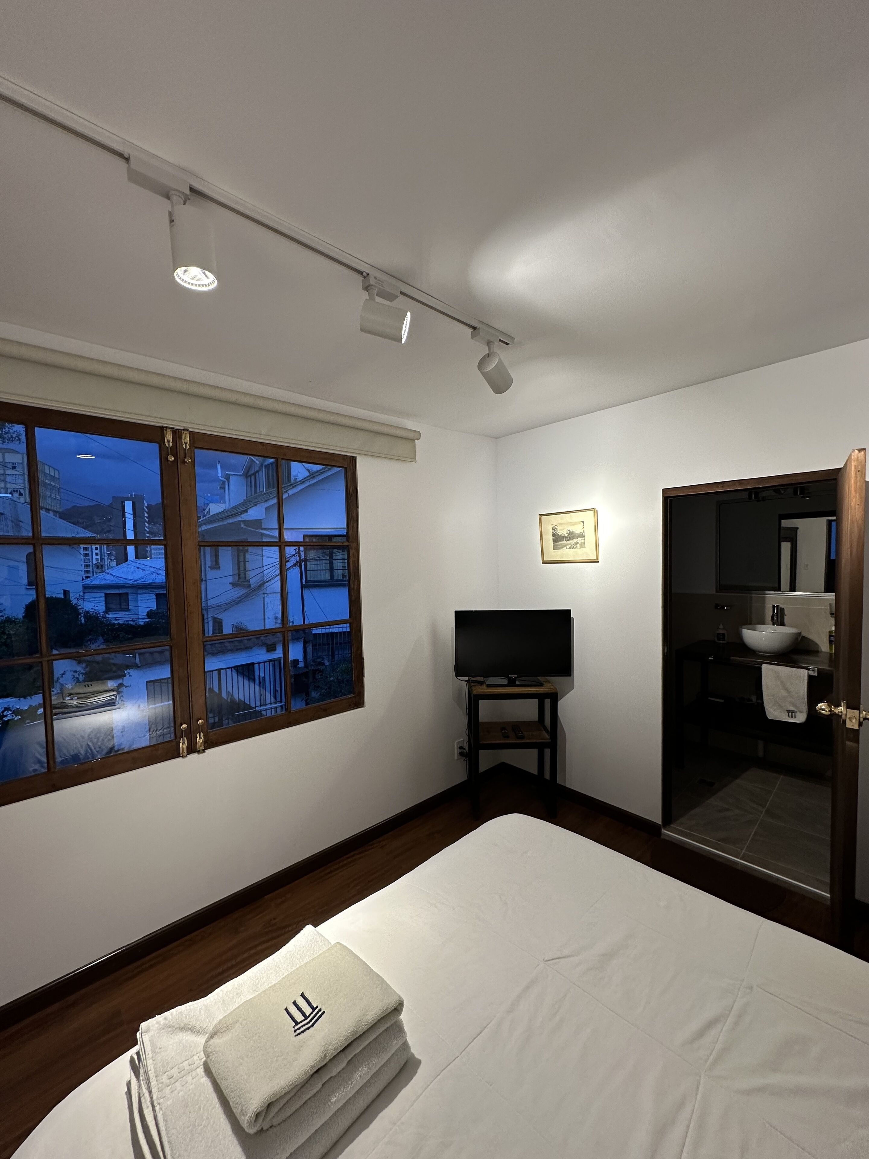 Una habitación de hotel con una cama, un televisor sobre un mueble, un baño con lavamanos y espejo, y una ventana con vistas a edificios.