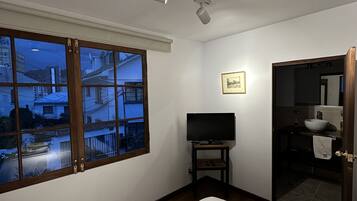 Una habitación de hotel con una cama, un televisor sobre un mueble, un baño con lavamanos y espejo, y una ventana con vistas a edificios.