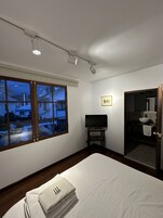 Una habitación de hotel con una cama, un televisor sobre un mueble, un baño con lavamanos y espejo, y una ventana con vistas a edificios.