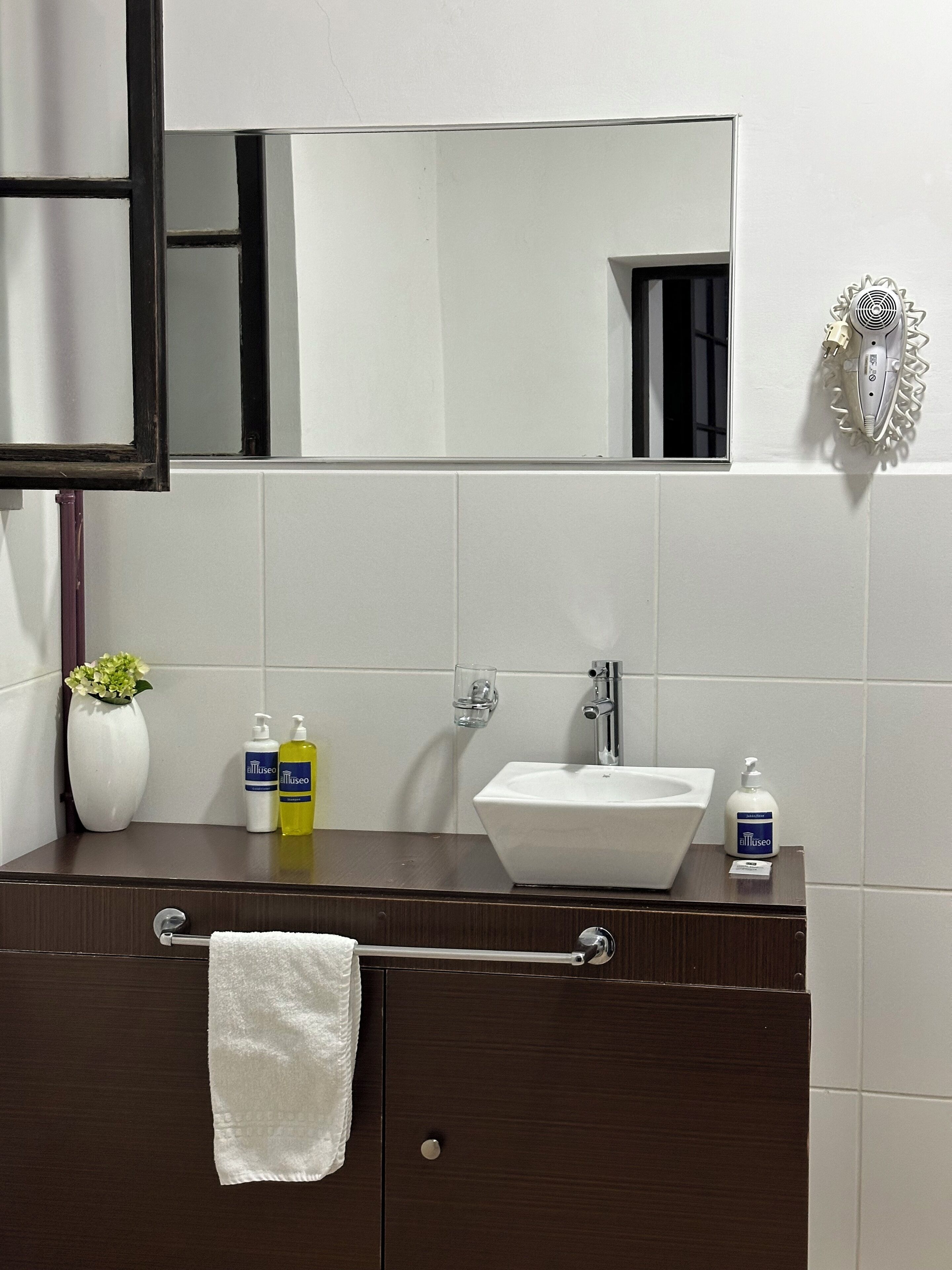 Un baño con tocador de madera oscura, un lavamanos blanco y un espejo encima. Hay artículos de aseo en el tocador, incluyendo una botella de jabón para manos y un vaso de agua.