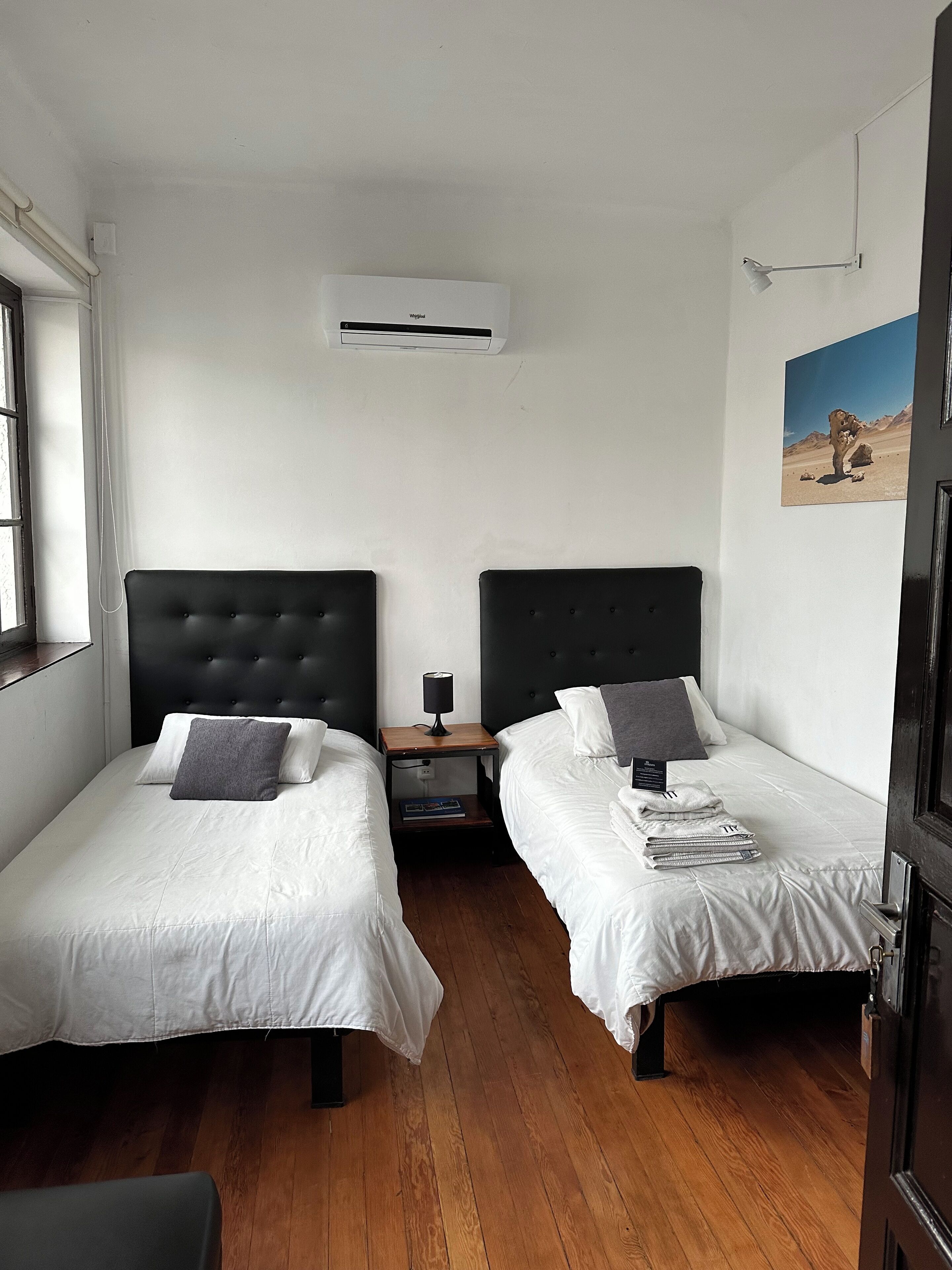 A hotel room with two single beds, a wooden floor, a white air conditioning unit, a nightstand with a lamp, and a painting on the wall.