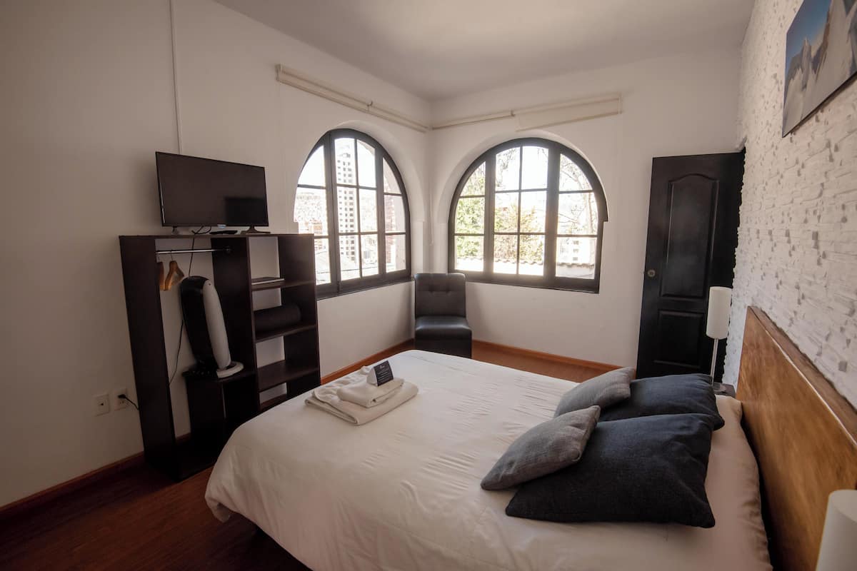 A hotel room with a bed, a TV, a wardrobe, and two windows with arched tops.