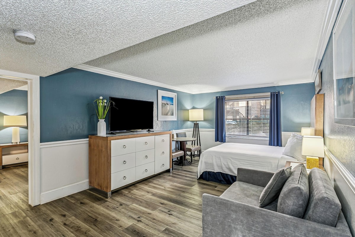 A hotel room with a bed, a sofa, a TV, a desk, and a window with curtains.