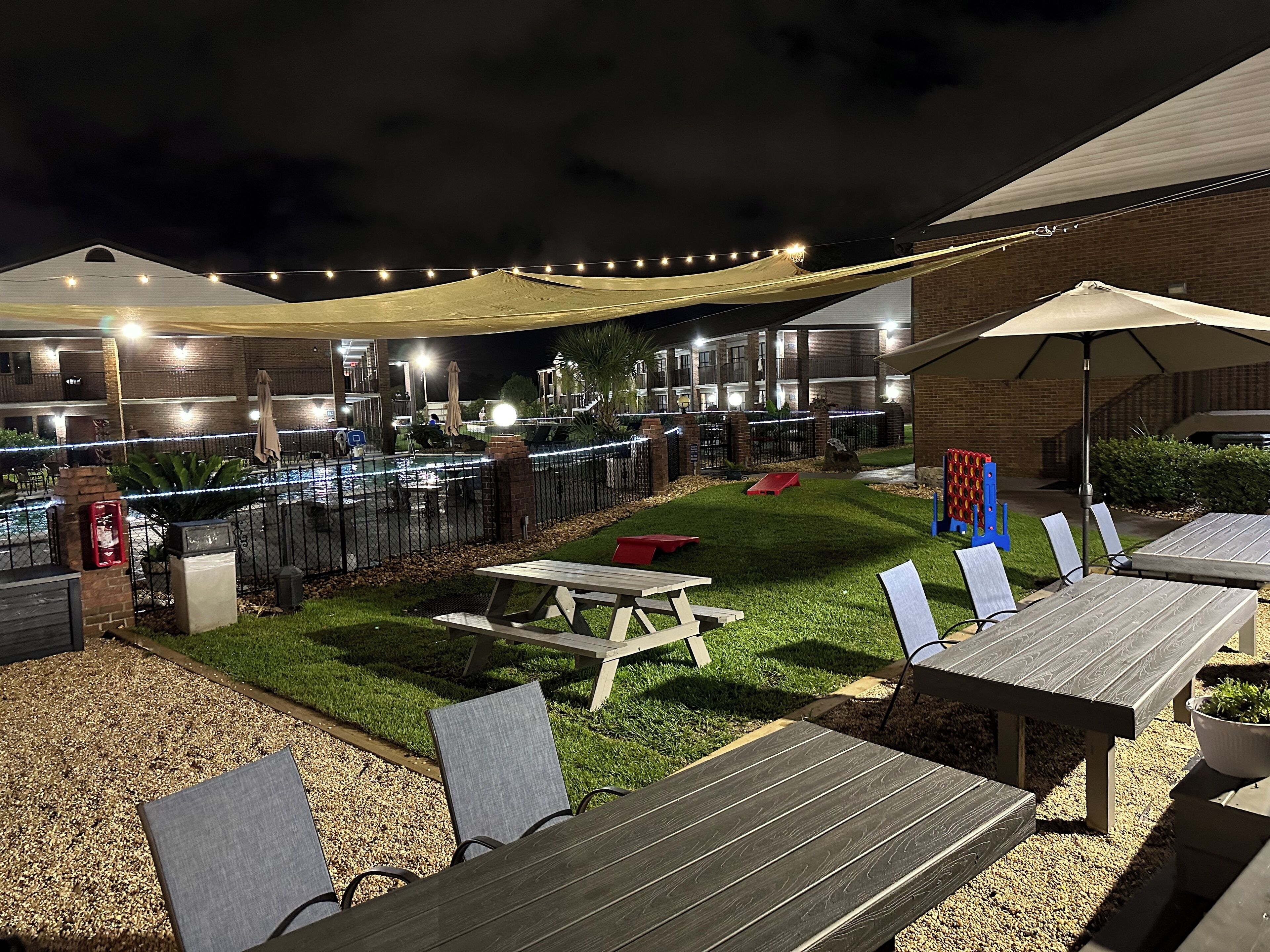 Un patio al aire libre con mesas y sillas de madera, un pequeño césped y luces colgantes.