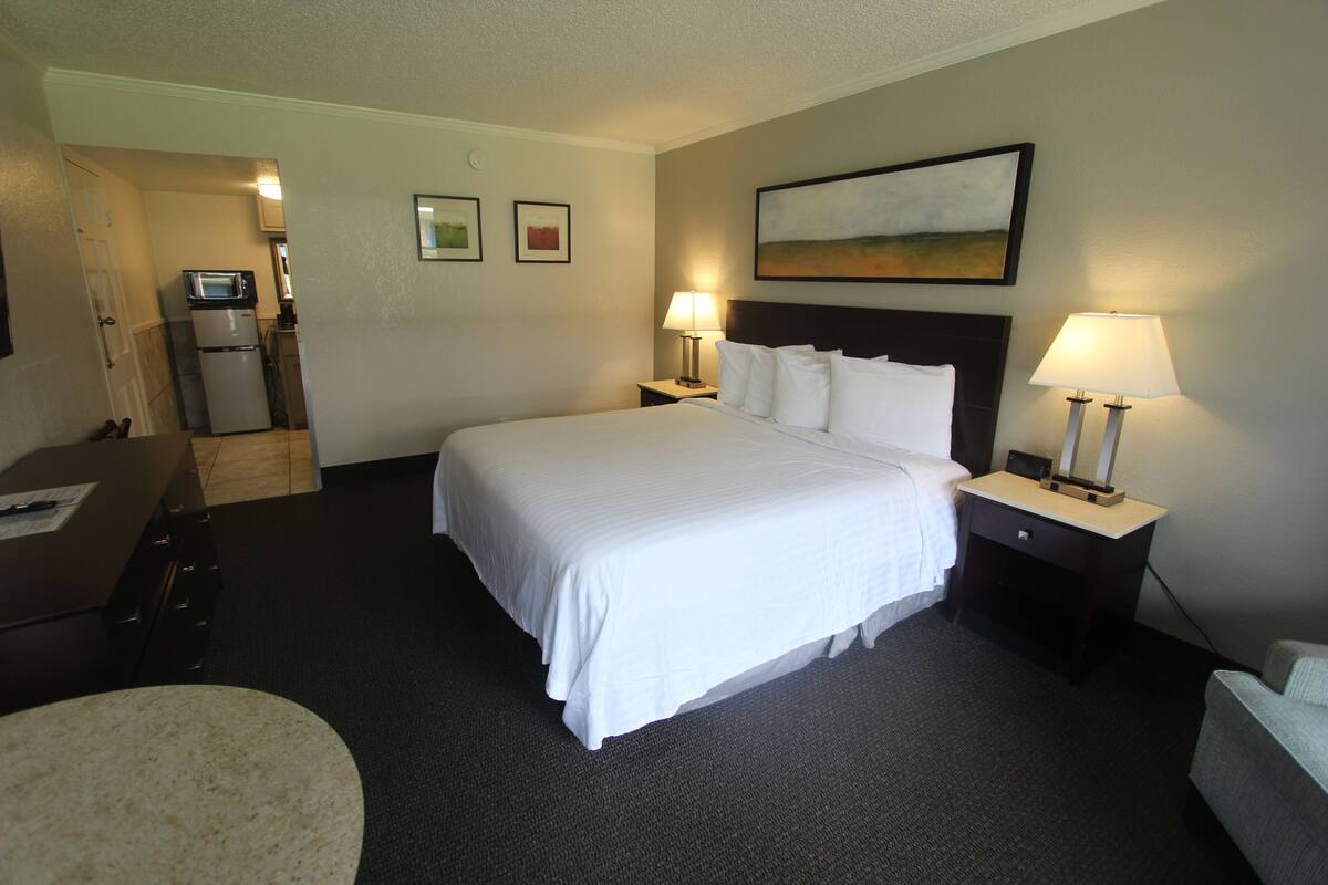 A hotel room with a large bed, two bedside tables with lamps, a desk, a chair, a microwave, and a refrigerator.