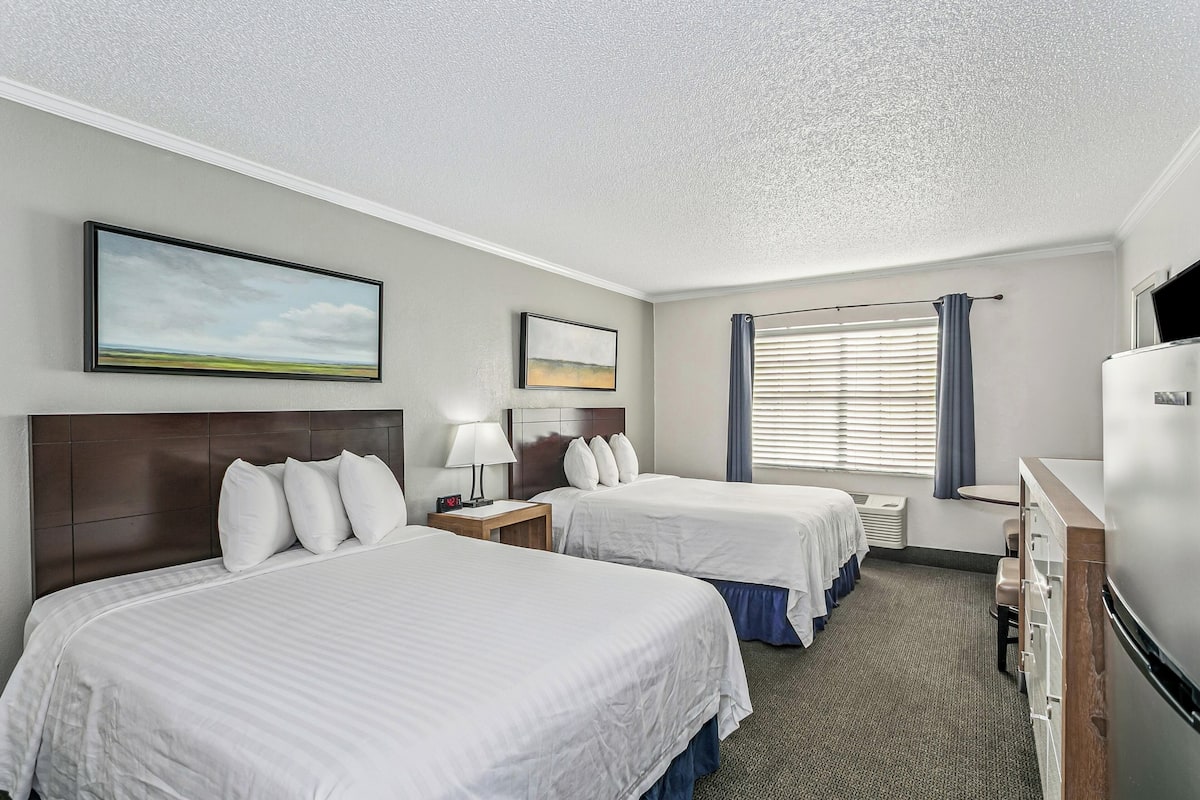 A hotel room with two beds, a desk, a refrigerator, and a window with blinds.