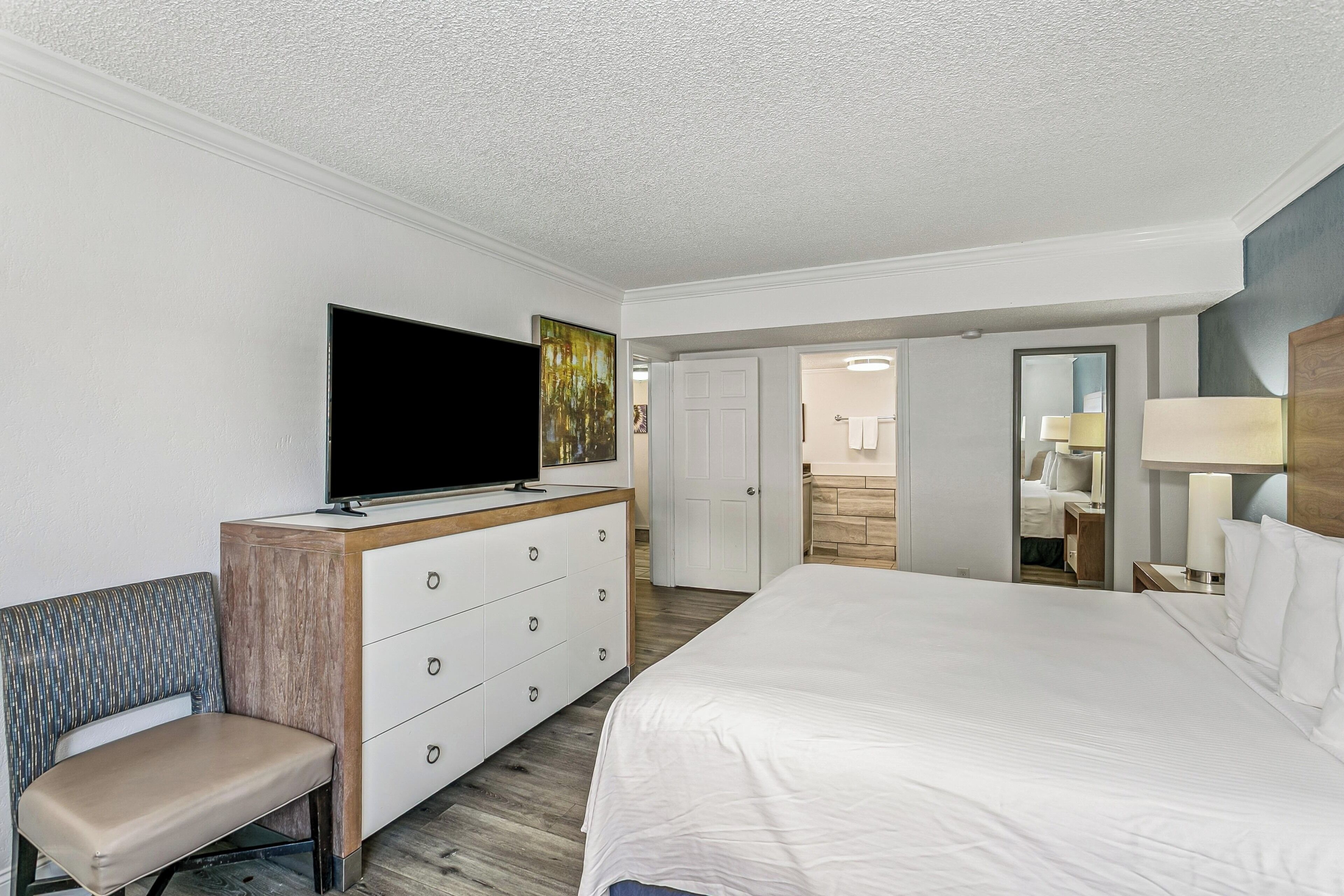 A hotel room with a bed, a TV, a dresser, a bench, and a bathroom visible through an open door.
