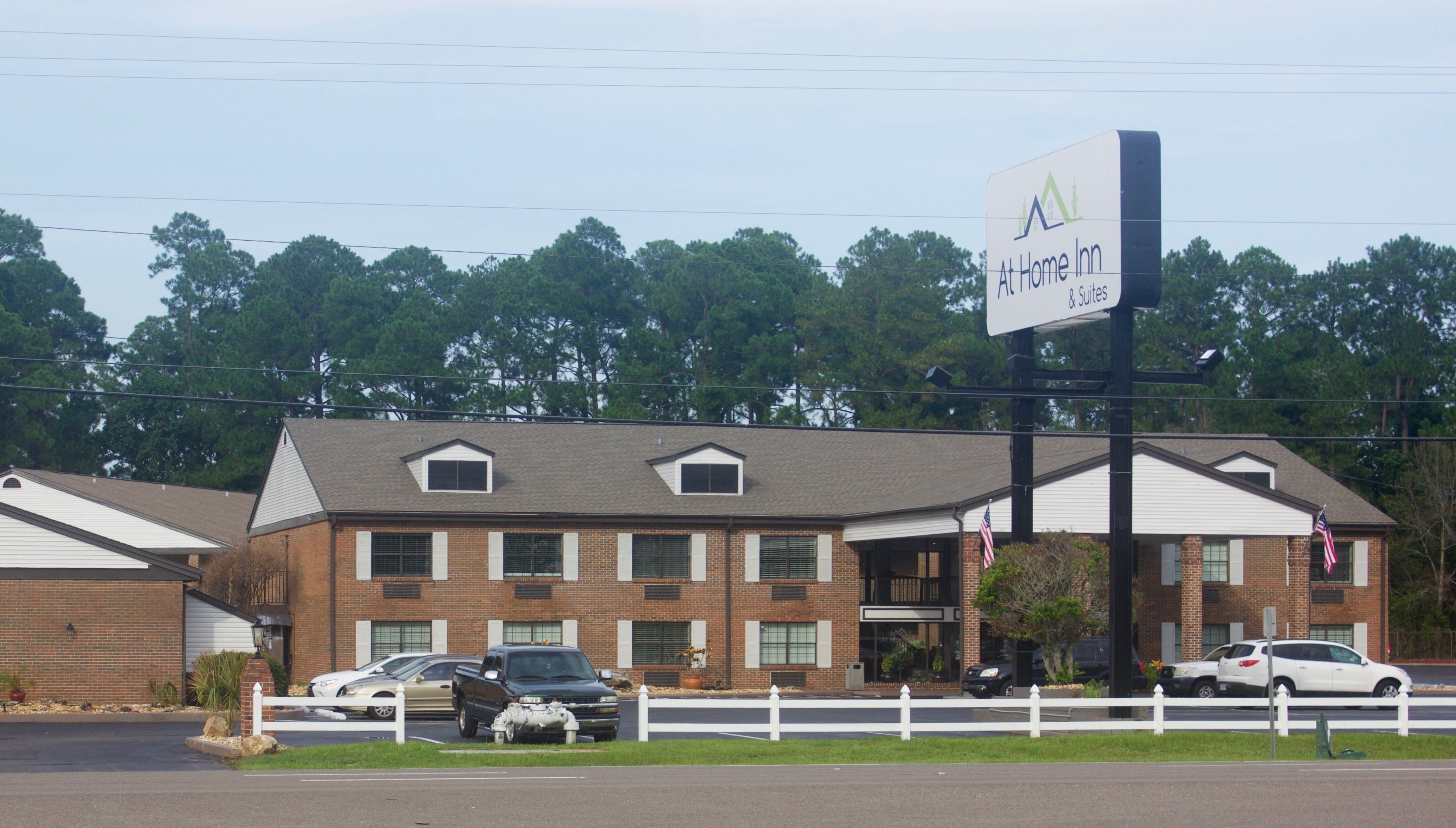 En toetages murbygning med et skilt, der viser 'At Home Inn & Suites'. Der er biler parkeret foran, et hvidt plankeværk og et flagstang med et amerikansk flag.