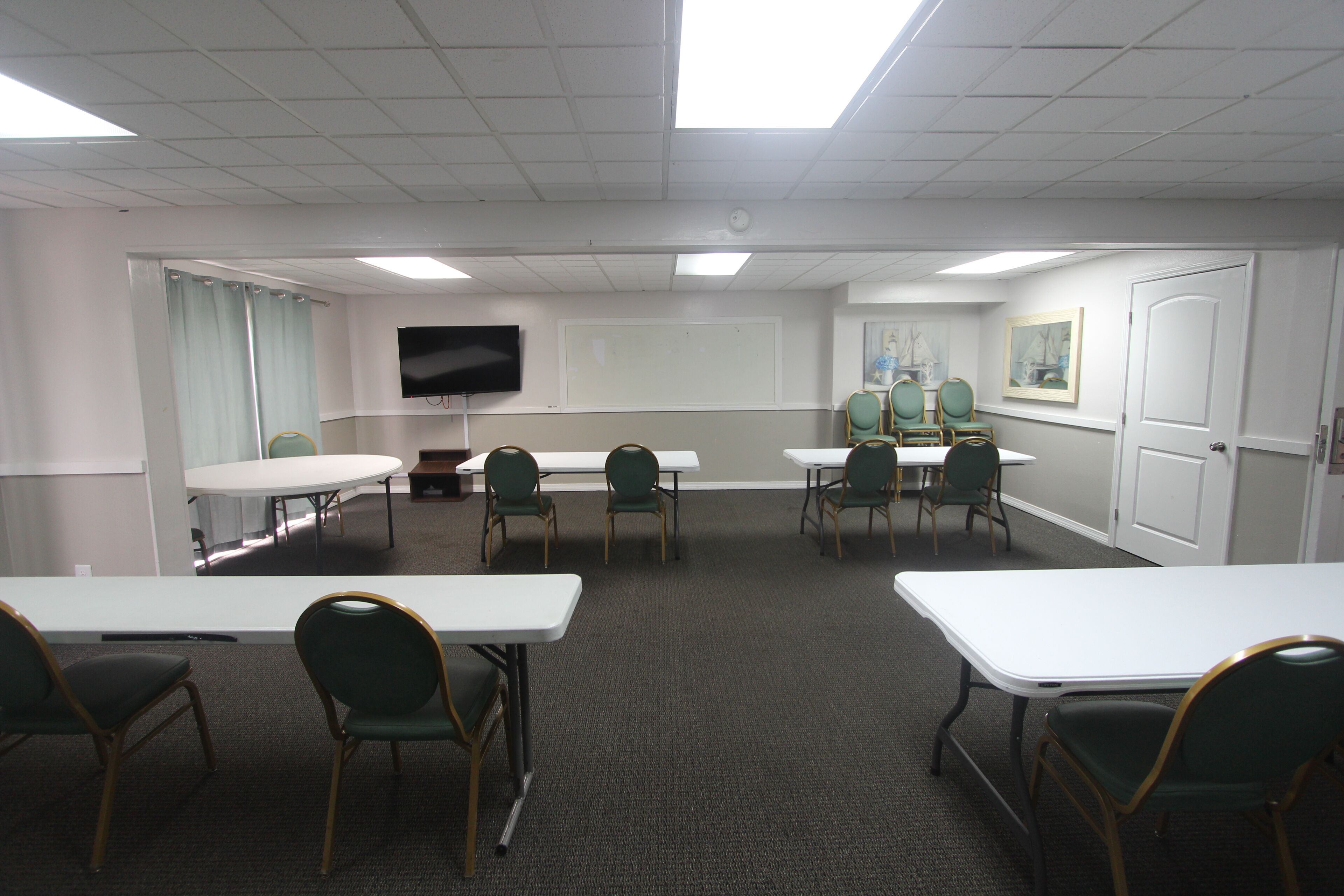 Un salón de conferencias con mesas blancas y sillas verdes, una televisión de pantalla plana y una pizarra.