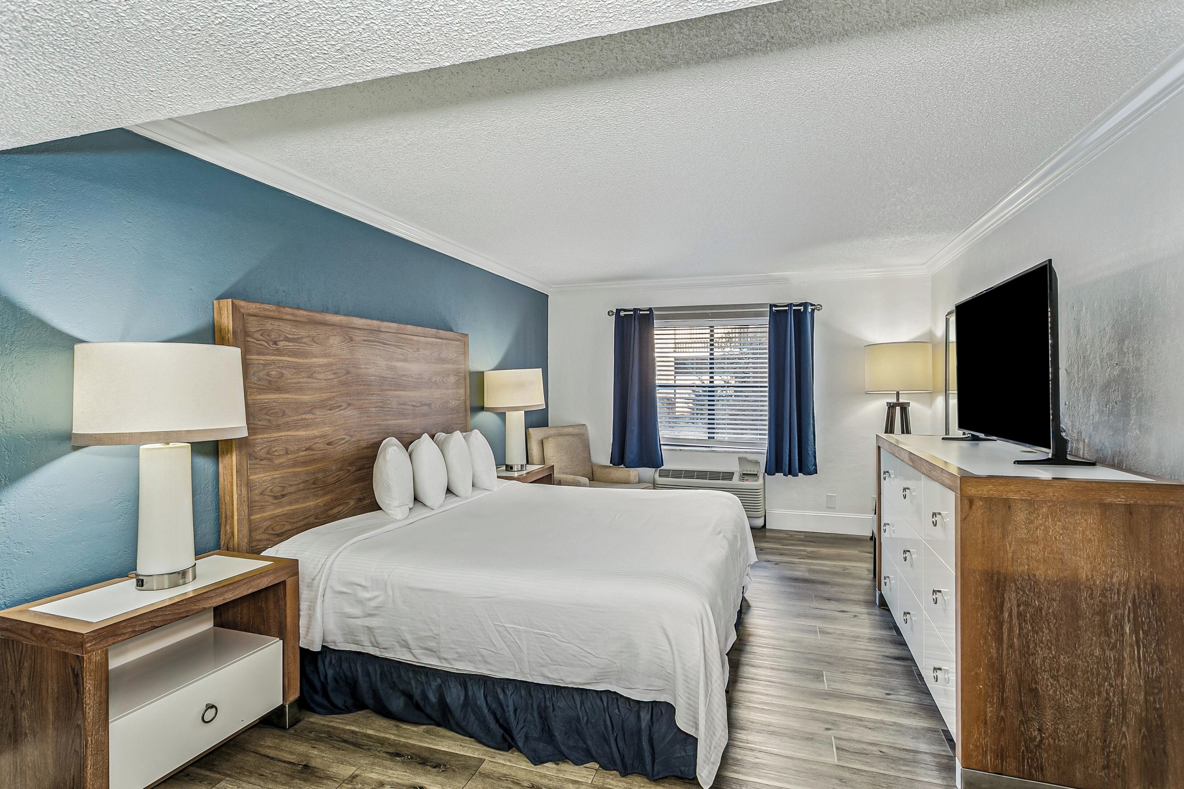 A hotel room with a large bed, a wooden headboard, a TV, a desk with a lamp, and a dresser.