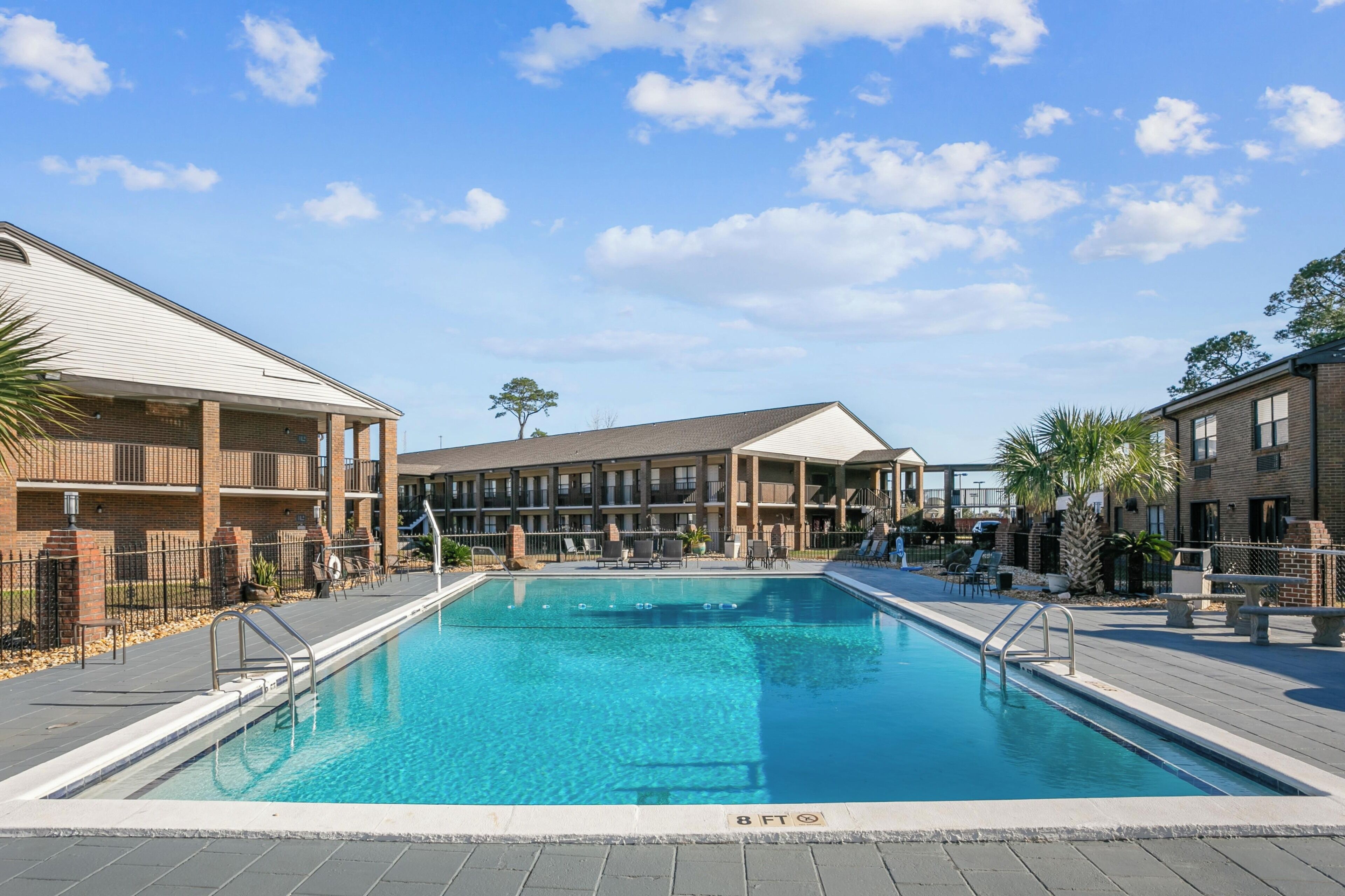 A swimming pool surrounded by multi-story buildings with balconies, outdoor seating, and a clear sky.