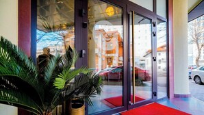 Entrada de hotel con puerta de vidrio, felpudo rojo y una planta de palma.