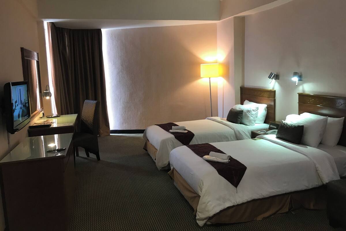 Une chambre d’hôtel avec deux lits, un bureau avec une télévision, une lampe et une vue sur l’extérieur.
