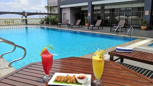A poolside table with two drinks and a plate of snacks, surrounded by lounge chairs and a building with large windows.
