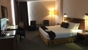A hotel room with a bed, a desk with a computer, two chairs, a lamp, and a TV.